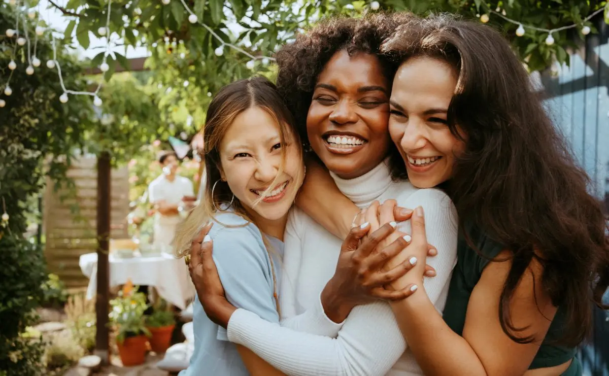 Three women smiling and hugging outdoors in a garden setting